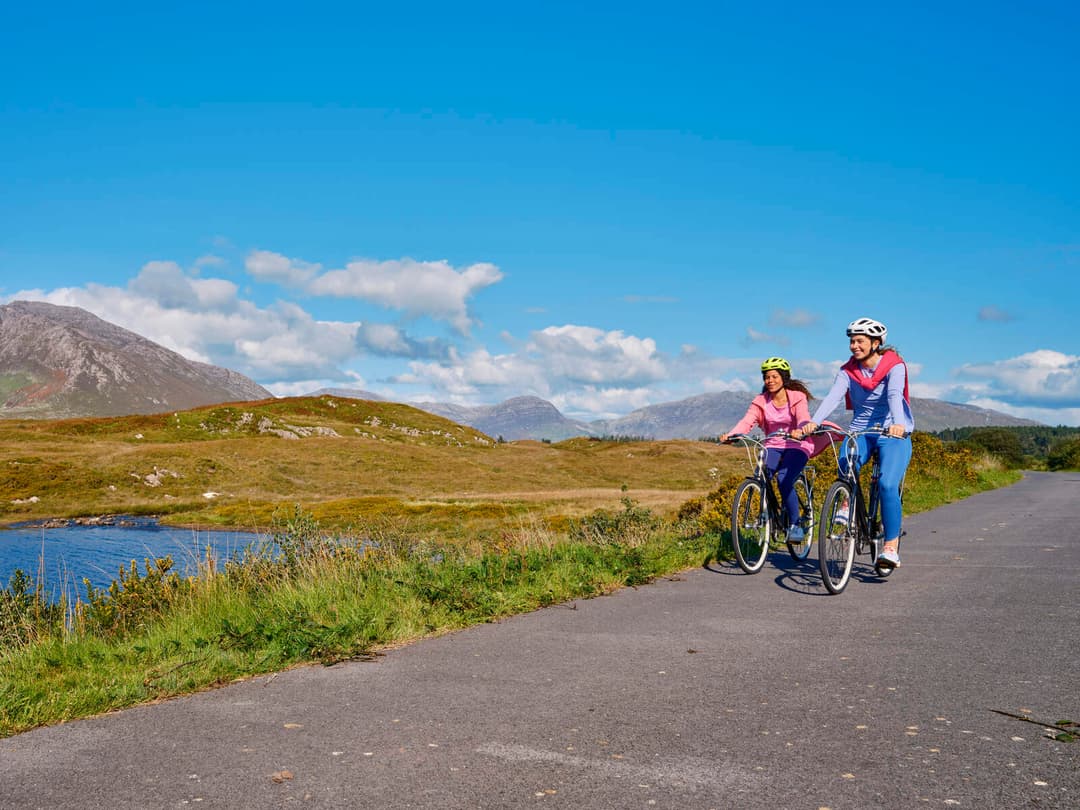 Two women cycling along a quiet Connemara road with mountains and a lake behind